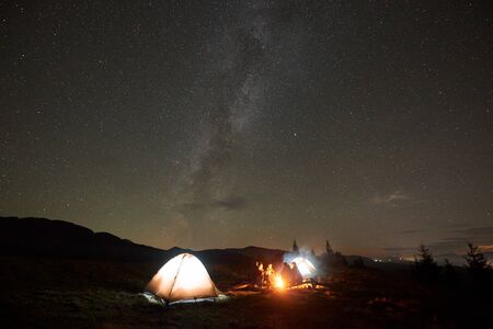 Night camping under stars. Group of five tourists, men and woman with guitar sitting by burning bonfire at tourist tents under dark sky with bright sparkling stars and Milky Way constellation.の写真素材