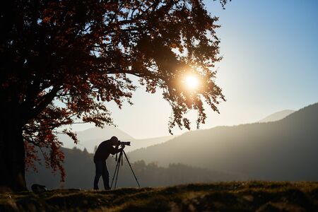 Tourist photographer using tripod and professional camera to take picture of mountain panorama, standing under large oak tree on woody foggy mountains landscape and blue sky at sunset background.の写真素材