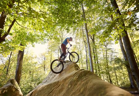Athlete male cyclist balancing on trial bicycle, biker making acrobatic stunt on big boulder in the forest outdoor on summer sunny day. Concept of extreme dangerous sport active lifestyleの写真素材