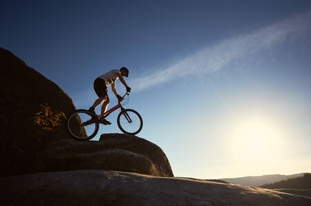 Silhouette of man cyclist balancing on trial bicycle, making acrobatic trick on top of mountain on summer sunny evening, blue sky and sunset on background. Concept of extreme sport active lifestyleの写真素材
