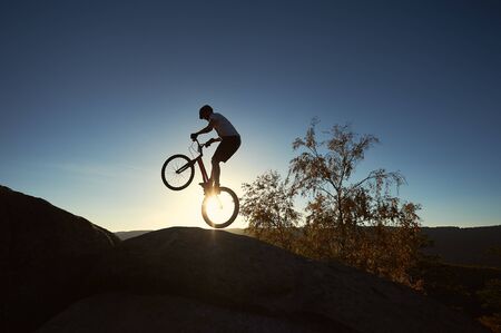 Silhouette of young cyclist balancing on back wheel on trial bicycle. Sportsman biker making trick on the edge of big rock on the top of mountain at sunset. Concept of extreme sport active lifestyleの写真素材