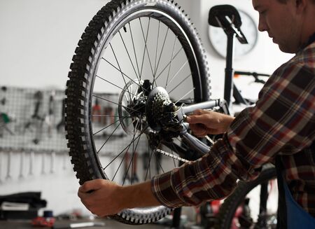 Cropped shot of male mechanic working in bicycle repair shop, repairman fixing bike using special tool, wearing protective workwearの写真素材