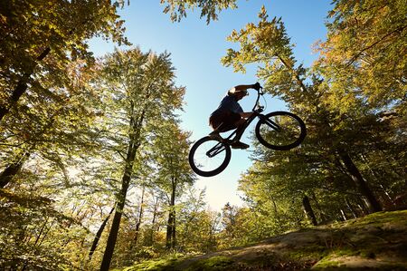 Young male bicyclist jumping on trial bicycle on big boulder, professional sportsman making acrobatic stunt in the forest on sunny day. Concept of extreme sport active lifestyleの写真素材