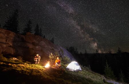 Night camping in the mountains. Happy couple travellers sitting together beside campfire and glowing tourist tent. On background big boulder, forest and night starry sky full of stars and Milky way.の写真素材