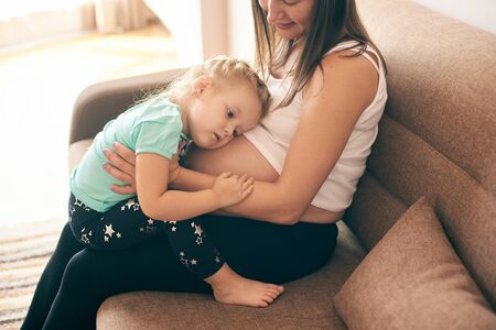 Side view of cute girl embracing big belly of pregnant mother. Happy woman and child sitting together on sofa and expecting little baby. Concept of parental love and pregnancy.の写真素材
