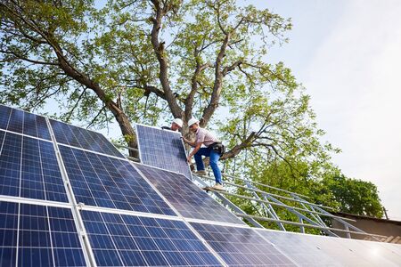 Two technicians mounting heavy solar photo voltaic panel on tall steel platform on green tree and blue sky background. Exterior solar panel voltaic system installation, dangerous job concept.の写真素材