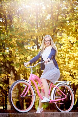 Active blond long-haired attractive traveler girl in glasses, skirt and blouse posing on modern pink lady bicycle on lit by autumn sun park stairs on bright colorful golden bokeh trees background.の写真素材