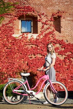 Young pretty long-haired smiling blond woman in casual clothing and glasses posing at pink lady bicycle on bright warm sunny day, on background of brick wall overgrown with beautiful red ivy leaves.の写真素材