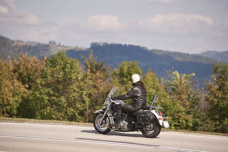 Side view of handsome bearded motorcyclist in helmet, sunglasses and black leather clothing riding cruiser motorbike along narrow asphalt path on sunny summer day.の写真素材