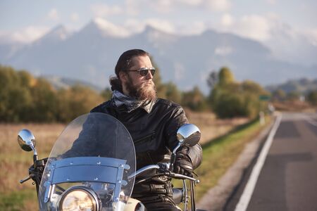 Portrait of handsome bearded biker in black leather jacket holding motorcycle handles on country roadside on blurred background of green landscape, distant white mountain peaks and bright sky.の写真素材