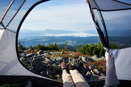 Camping on the top of mountain on summer morning. Young hiker woman sitting in the entrance of tourist tent. On foggy mountains background. View of legs. Point of view shot. Tourism adventure conceptの写真素材