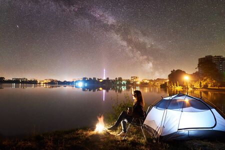 Night camping on lake shore near campfire. Woman with cup of coffee sitting on chair near tourist tent, enjoying evening sky full of stars and Milky way, quiet water surface, city lights on backgroundの写真素材