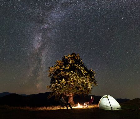 Romantic couple hikers enjoying summer night camping in mountains. Man and woman sitting on chairs beside bonfire, glowing tourist tent and big tree under starry sky full of stars and Milky way.の写真素材