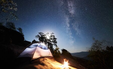 Tourist camping at summer night on rocky mountain. Illuminated tent and campfire under wonderful night sky full of stars and Milky way. On the background beautiful starry sky, big boulders and treesの写真素材