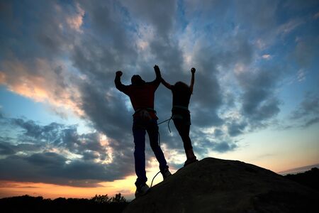 Teamwork couple helping hand trust help, silhouette success in mountains. Team of climbers man and woman. Hikers celebrate with hands up, help each other on top of mountain, happy climbing together.の写真素材