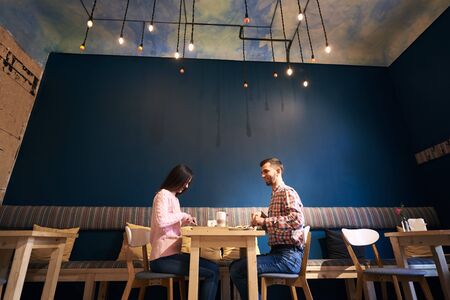Young couple sitting in atmospheric cafe with interesting ceiling by dating indoors. Full length profile shot of a male and female student having dinner and talking in cafe isolated on blue backgroundの写真素材