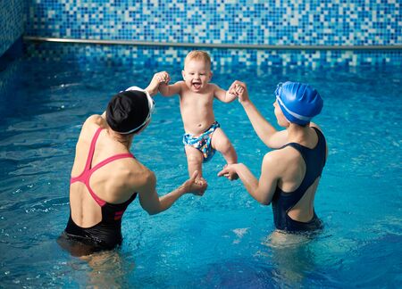 Back view of two women holding babys hands and feet, rising up in air laughing boy from water after swimming in pool. Amusement for baby at swimming classes. Having fun together. Sports and familyの写真素材