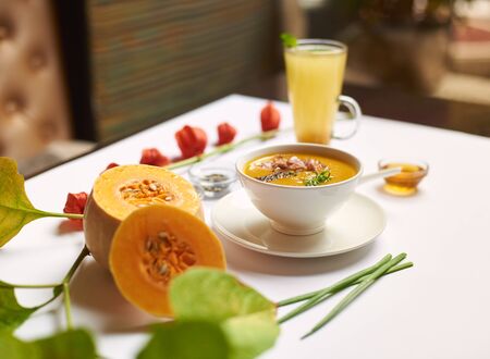 Creative still life served on the table for dinner. Delicate cream soup and tea in high cup prepared in luxury restaurant. Green leaves and halves of pumpkin lying near meal on white tablecloth.の写真素材