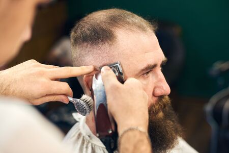 Side view portrait of adult dark-haired bearded man in modern barbershop during haircutting process with electric razor. Professional tools and accessories in barbers hands. Blurred dark background.の写真素材