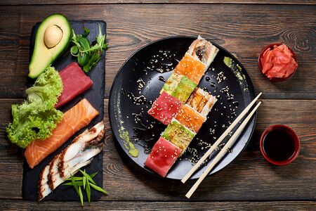 Delicious sushi with rice and fish on black plate served on the wooden table, decorated with chopsticks, bowl of soy sauce, avocado and sprouts. View from aboveの写真素材