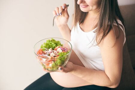View from above of cheerful pregnant woman in shirt resting at home and eating healthy food. Smiling woman keeping big plate with vegetables. Concept of healthy lifestyle.の写真素材