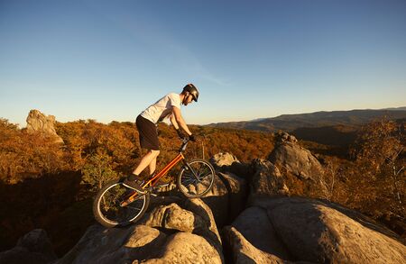 Professional cyclist balancing on trial bicycle on top of big rock, male biker making acrobatic stunt on summer evening, blue sky and forest on background. Concept of extreme sport active lifestyleの写真素材