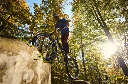Low angle view of male cyclist balancing on trial bicycle, making acrobatic stunt on big boulder in the forest on summer sunny day. Concept of extreme dangerous sport active lifestyleの写真素材