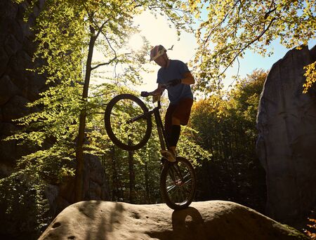 Professional sportsman bicyclist balancing on back wheel on trial bicycle, male cyclist making acrobatic stunt on big boulder in the forest on sunny day. Concept of extreme sport active lifestyleの写真素材