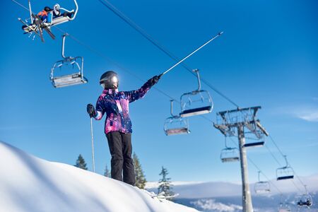 Low angle shot of female smiling skier standing on mountain slope and showing with ski poles something in distance on sunny winter day. Sunlight reflection in goggles. Active lifestyle at resort.の写真素材