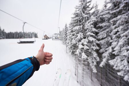 Point of view with males hand with thumb up for snowy nature forest around ski lift. Unrecognizable man on ski lift. Cropped photo of skier hand, amazing monochrome mountain hill blurred view. POVの写真素材