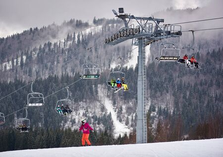 Bukovel, Ukraine - December 09, 2018: woman snowboarding down near cable chairlift line carrying passengers skiers and snowboarders up along snow-covered slope among wooded Carpathian mountainsのeditorial素材