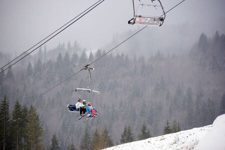 Bukovel, Ukraine - December 09, 2018: family three persons skiers with skis on their feet sitting on cable chairlift carrying up over dark coniferous forest covered with dense fog in winter mountainsのeditorial素材