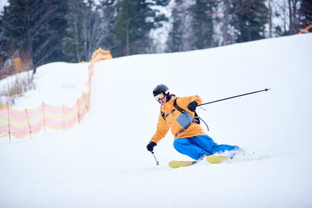 Front view of young skier concentrated on skiing down on steep ski slope. Proficient technical carving skiing. Net on slope edge. Wooded mountain top on background. Extreme winter activities conceptの写真素材