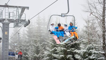 Bukovel, Ukraine - December 09, 2018: snowboarders men and women in ski mask sitting together in chairlift and carrying near huge steel pillar and along dense forest with fir trees in snowfallのeditorial素材