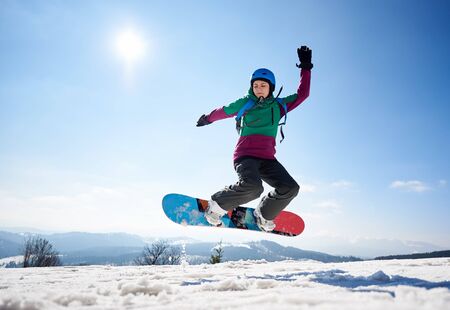 Young female snowboarder jumping in air on copy space background of clear bright blue sky and snowy mountains on sunny winter day. Winter sports and recreation, leisure outdoor activities concept.の写真素材