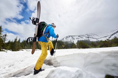 Back view of man carrying snowboard on back. Male extremal going on challenging snowy route to mountain top for off-piste snowboarding riding. Backcountry snowboarding tour popularization. Copy spaceの写真素材
