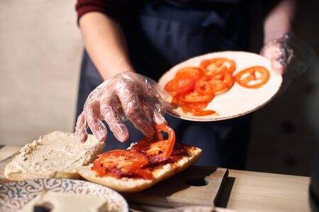 Chef mastery hands holding white plate with slices of cheese and sausage. Putting pieces of tomato on bread greased with red sauce for cooking panini. Wooden working surface. Blurred background.の写真素材