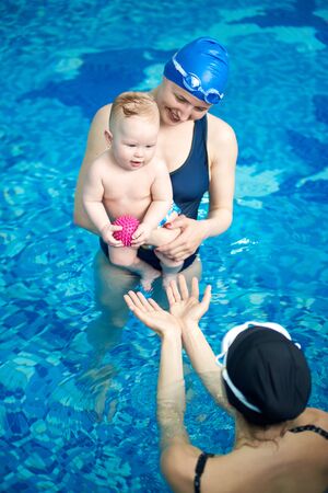 Top angle view of mom in swimsuit standing in swimming pool and holding baby her son in arms and laughing while another woman playing ball with her kid. Happy active healthy childhood conceptの写真素材