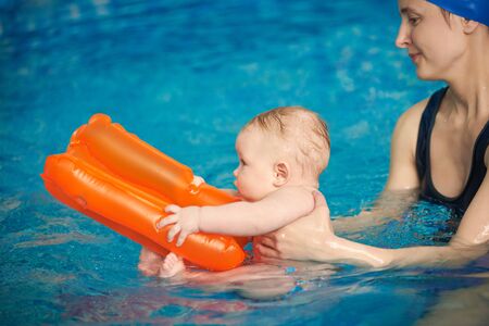 Inflatable orange toy helping little child staying on water and not sinking in swimming pool. Young beautiful woman bringing up new swimmer. Side view. Concept of benefits of swimming for infantsの写真素材