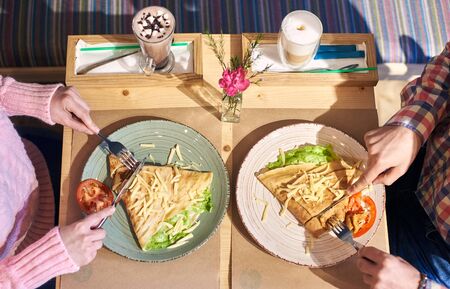 Cropped hands of man and woman savoring food omelet and coffee for lunch sitting at wooden table in cozy atmosphere. Healthy lunch served with vegetables and cheese. Beverages on background. Top view.の写真素材