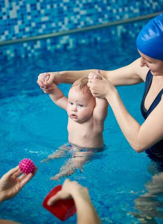 Concentrated active blond baby boy keeping upright in water. Cropped view of woman in swimwear holding child by his hands up, entertaining child with toys. Wellness classes for children in waterの写真素材