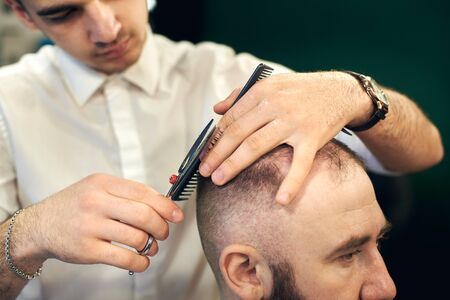 Cropped of male young hairdresser barber making short haircut for his client in modern barbershop. Concept of traditional haircutting with scissors. Step-by-step lesson for creating mens hairstyles.の写真素材