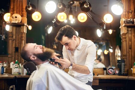 Young concentrated barber shaving and cutting beard with electric trimmer. Fashionable bearded lumberjack style. Barbershop interior with elements of eclecticism and creative lighting. Side view.の写真素材