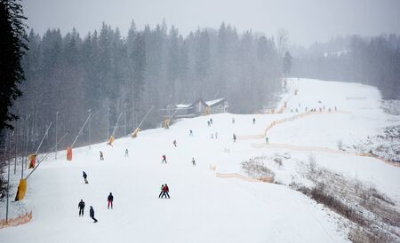 Bukovel, Ukraine - December 09, 2018: many people freeriding skiing and snowboarding on ski run with snow cannon along snowy cascading slope among mountains at ukrainian ski resort in snowfallのeditorial素材