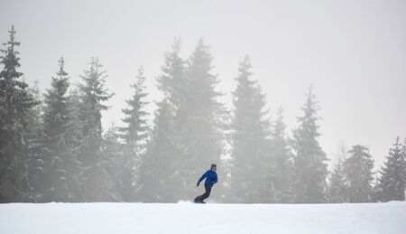 Sporty female snowboarder sliding on flat snow-covered road. Poor visibility in snowfall. Coniferous forest on background. Copy space. Monochrome view. Freedom, sport, winter activities conceptの写真素材