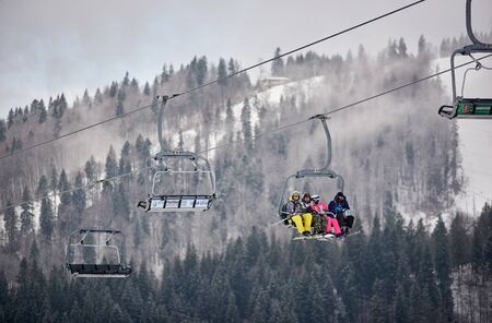 Bukovel, Ukraine - December 09, 2018: skiers and snowboarders sitting carrying high up on cable chairlift in one direction over snow-covered slopes in Carpathians mountains in foggy gray dayのeditorial素材