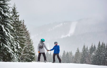 Young man and woman snowboarder standing on high mountain near fir trees along slope, holding hands and looking at each other. Snowfall in mountains. Picturesque winter landscape on background.の写真素材
