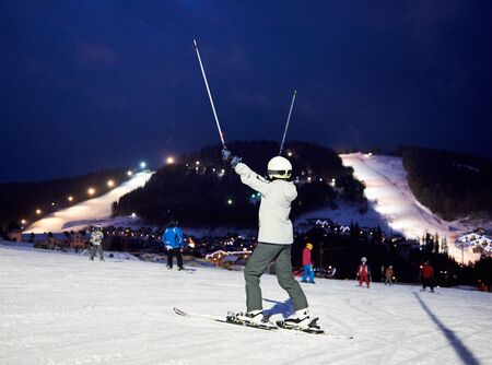 Back view of female skier rejoicing lifting hands up with ski poles. Happy ending of active sports skiing winter day at ski resort. On background mountains with ski slopes under dark blue sky.の写真素材