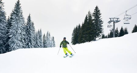 Male skier skiing on ski hillside at resort after taking chairlift on beautiful snow-covered mountain. Tourist backpacker freeriding on desert wooded slope. Ski chairlift and grey sky on background.の写真素材