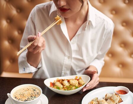 Crop of pretty woman in white blouse with red lips ordering meal in luxury Japanese restaurant. Girl eating four course lunch including soup, tempura shrimps, noodles and warm salad.の写真素材